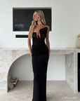 A blonde woman models a strapless black evening gown with a sheer lace corset-style bodice and a fitted floor-length skirt, standing in front of a modern white wall and marble fireplace.